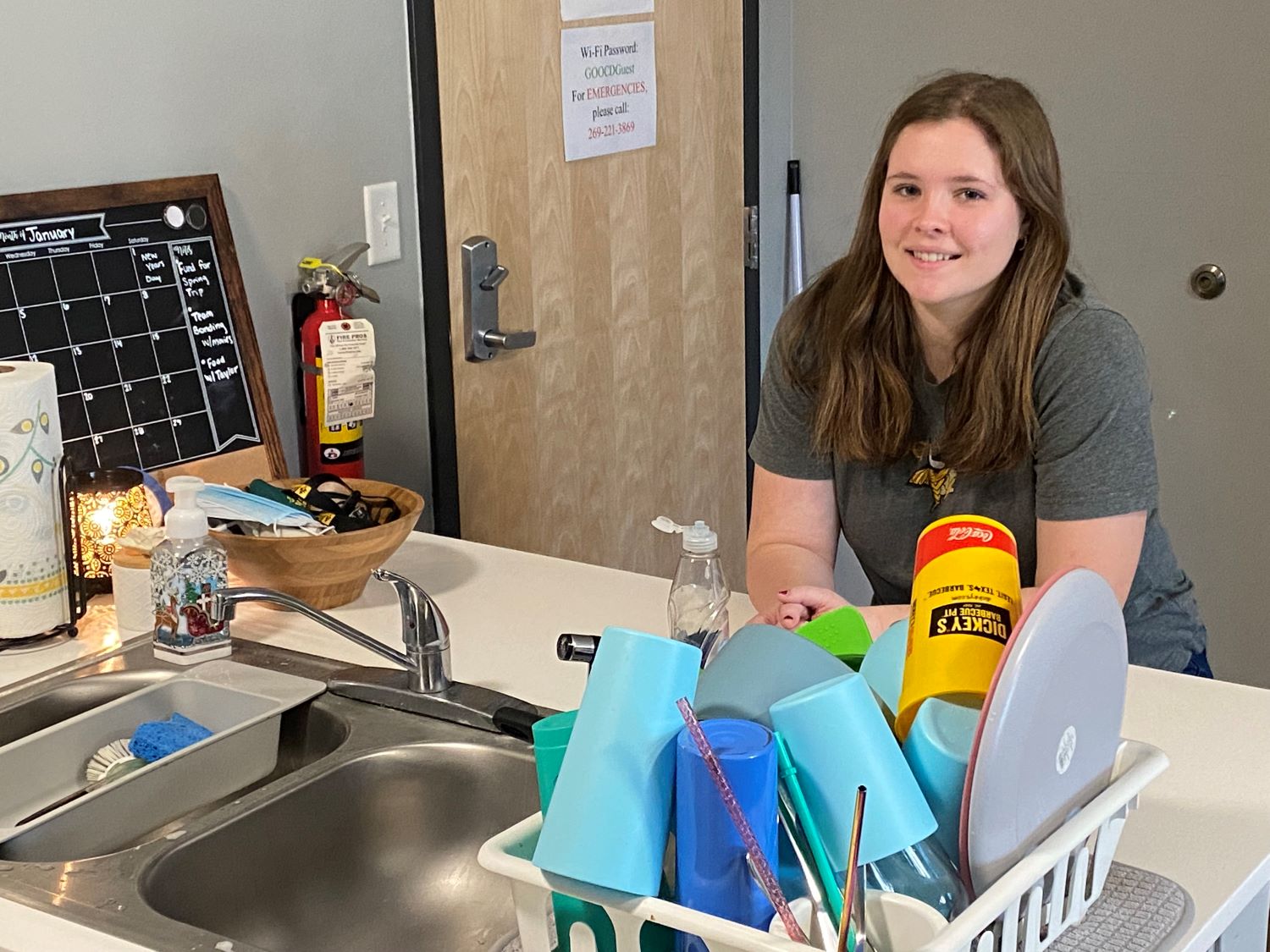 Female student leaning on her kitchen counter in front of her sink with clean dishes stacked on the counter, smiling at the camera.