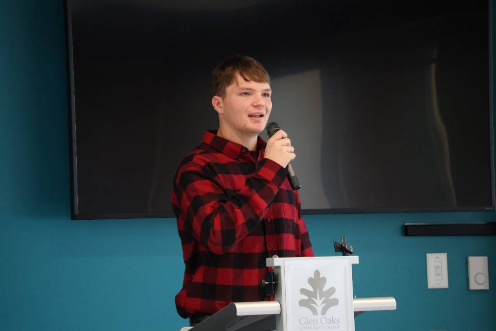 male student in lumberjack shirt, holding microphone and speaking for communications presentation