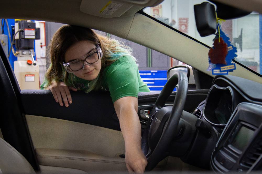 Female student reaching inside of the interior of a car from outside of the window.