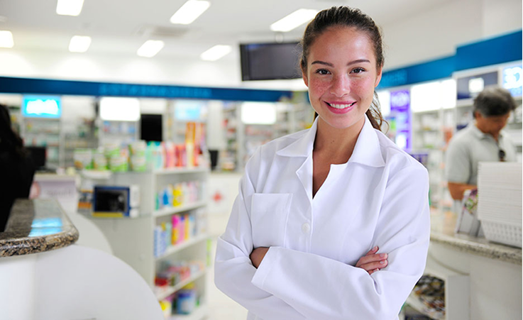 Female in a white pharmacy coat, standing with her arms crossed and smiling in the pharmacy area
