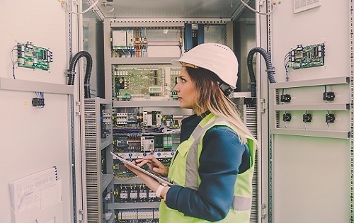 Female electrician standing in a hard hat, wearing a vest, in an electrical room